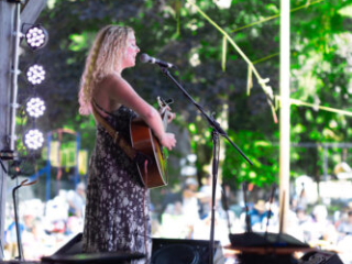 A photo of an artist singing into a mic and playing acoustic guitar, taken from the side with a background of trees